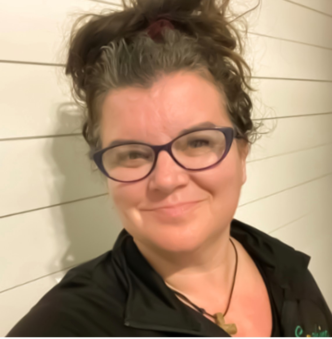 Haley Hennlich is a white woman with glasses and curly hair pulled back into an updo. She is wearing a dark shirt and standing in front of a white shiplap background.